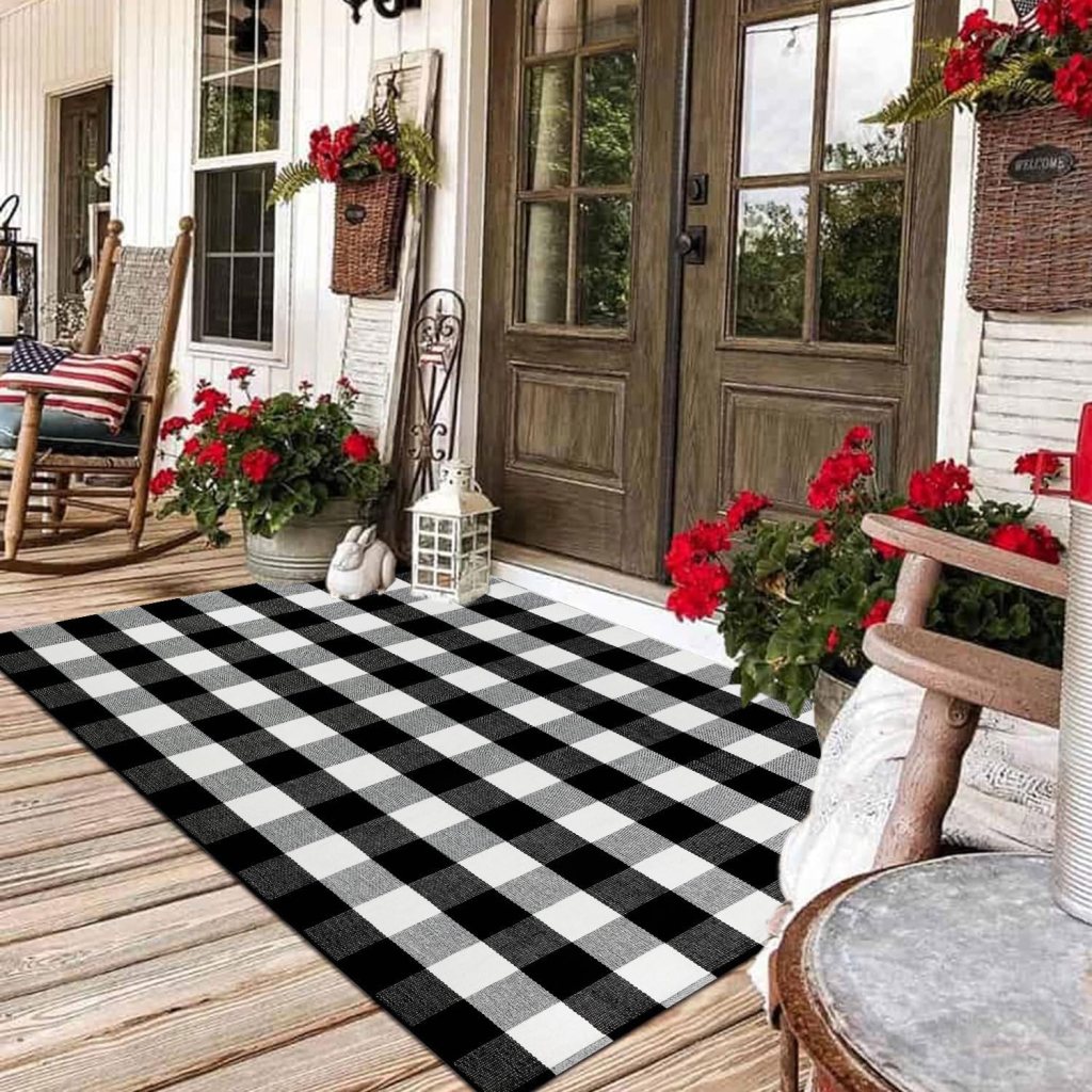 A cozy porch entrance featuring a black and white checkered rug, rocking chairs, blooming red flowers in pots, and a lantern beside double doors.
