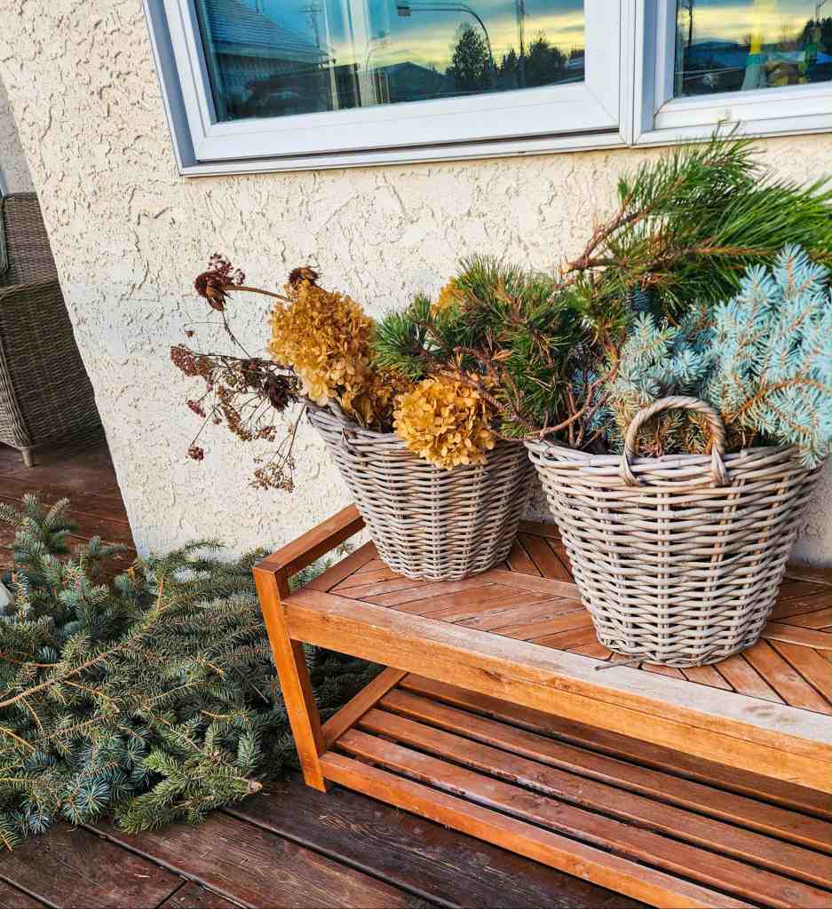 Two wicker baskets filled with dried flowers and evergreen branches on a wooden bench outside a house.