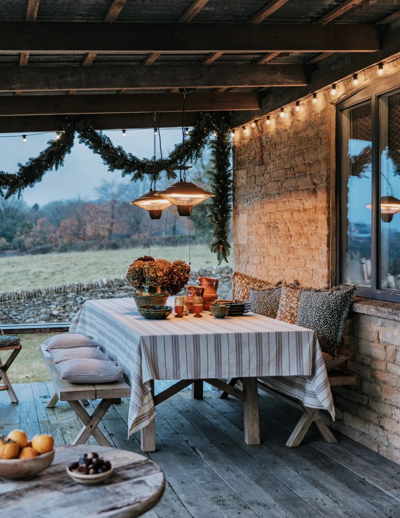 A cozy outdoor dining space featuring a striped tablecloth, rustic wood furniture, and decorative items like potted flowers, glasses, and plates. The area is adorned with garlands and string lights, creating a warm ambiance against a backdrop of a scenic landscape.