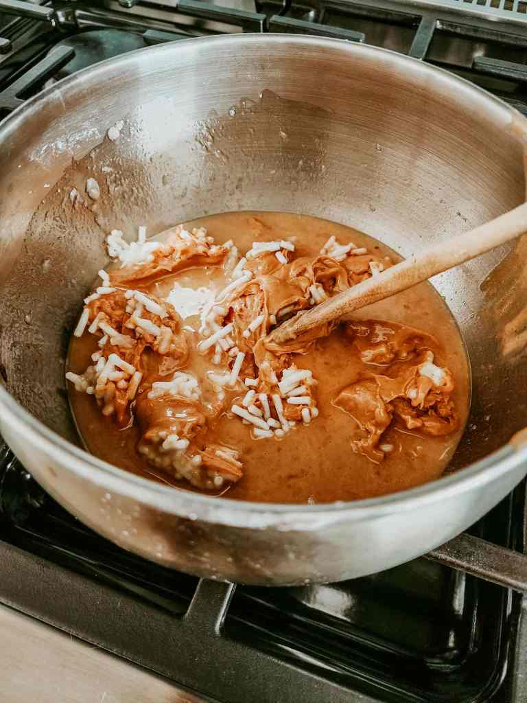 A large stainless steel mixing bowl on a stove containing melted peanut butter and suet, with chunks of brown ingredients and white rice-like items being stirred with a wooden spoon.