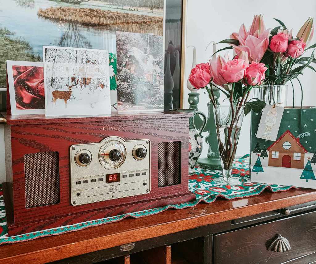 A vintage-style radio on a wooden side table, decorated with holiday greeting cards and a bouquet of pink roses in a vase. The table is covered with a festive tablecloth.