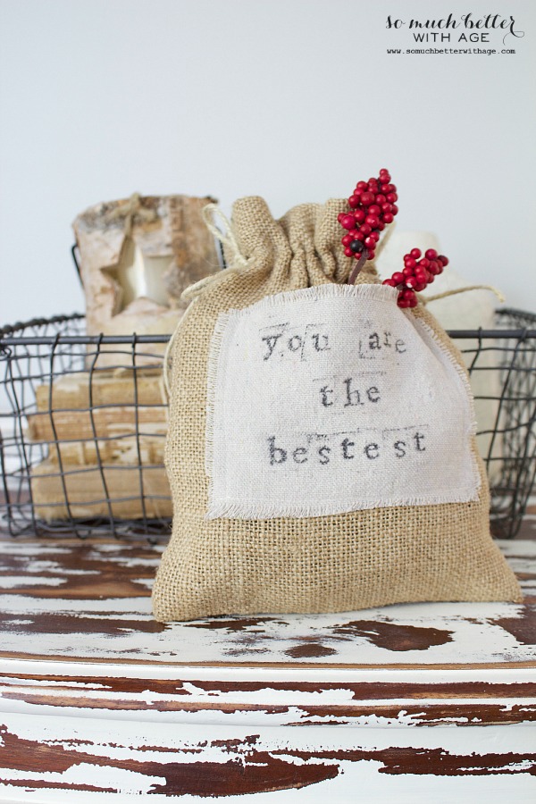 A decorative burlap bag with the printed message 'You are the bestest,' adorned with red berries, placed on a rustic wooden surface next to a wire basket containing wooden blocks.