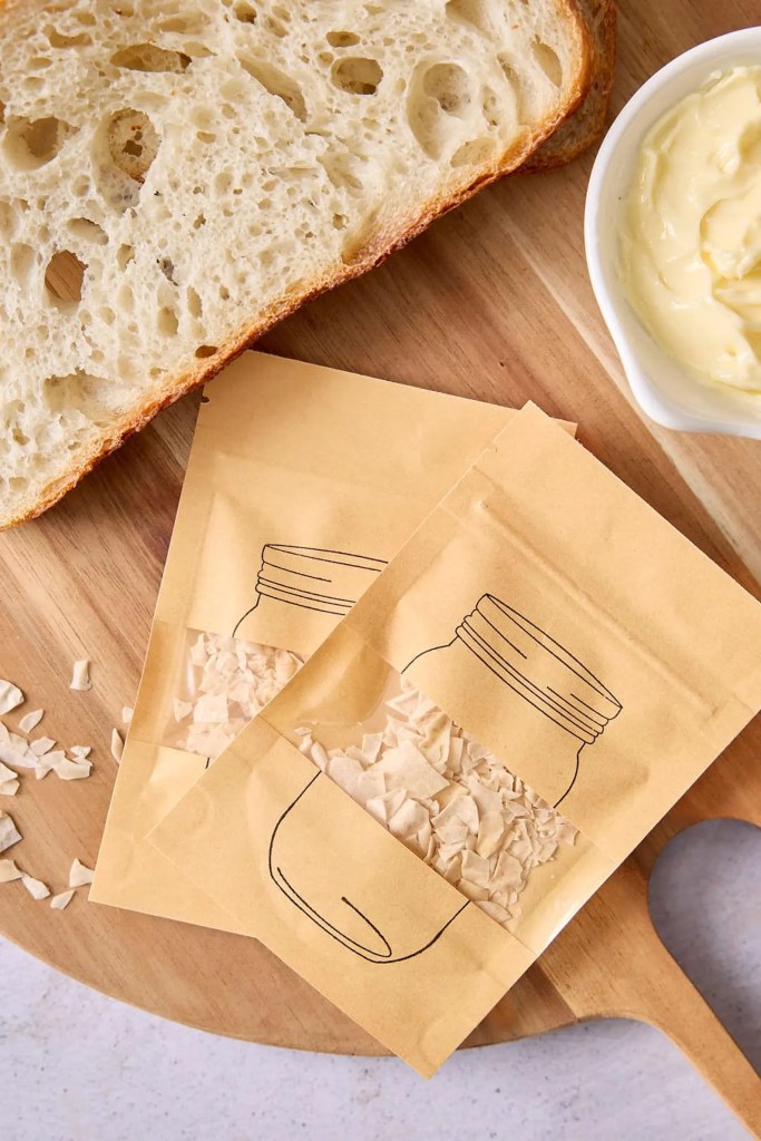 Sliced artisan bread on a wooden cutting board with two decorative brown paper packets resembling jars filled with flakes, alongside a small bowl of butter.