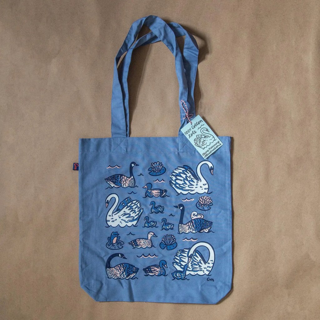 A blue cotton tote bag featuring whimsical illustrations of swans and water lilies, displayed against a brown background.