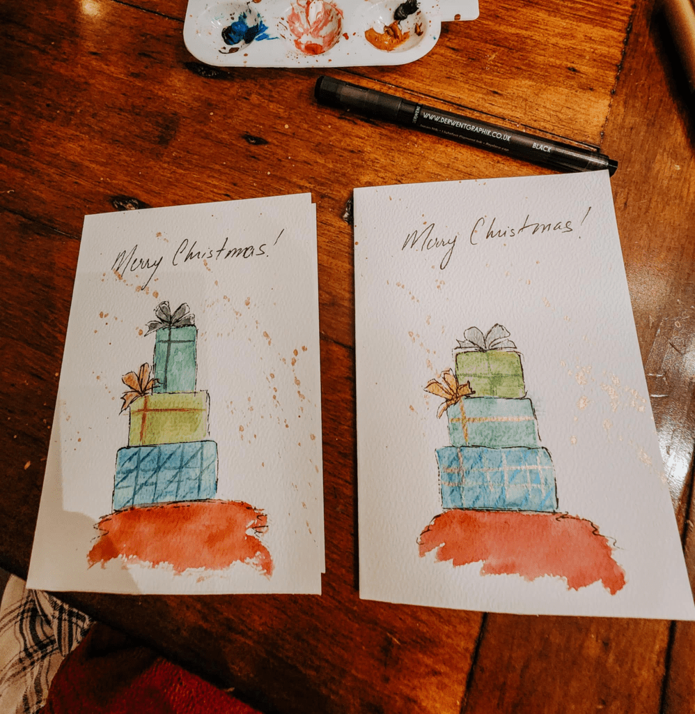 Two hand-painted Christmas cards featuring stacked presents with the message 'Merry Christmas!' at the top, placed on a wooden table with a palette of paints and a black pen.