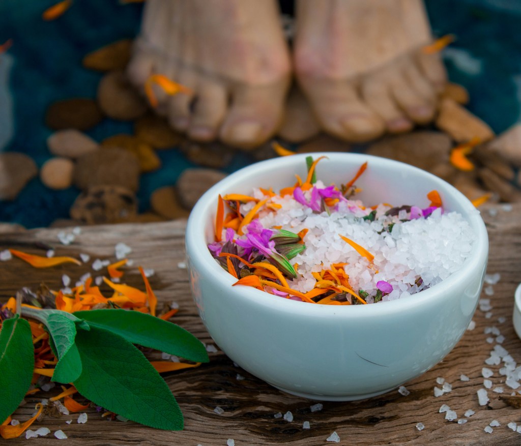 A close-up of a white bowl filled with bath salts and colorful flower petals on a wooden surface, with bare feet visible in the background.