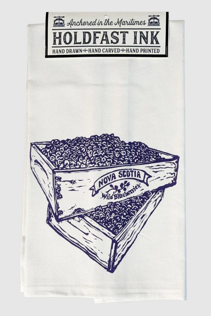 A white tea towel featuring a hand-drawn illustration of a wooden crate filled with blueberries, labeled 'Nova Scotia Wild Blueberries'.