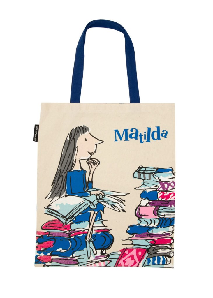 A tote bag featuring a whimsical illustration of a girl sitting on a pile of books, with the name 'Matilda' printed in blue.