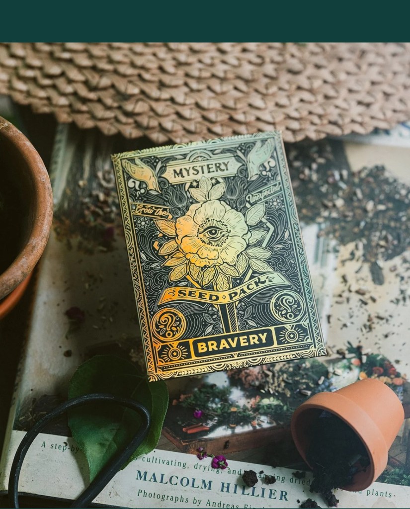 A decorative seed pack labeled 'Bravery' with intricate designs, placed next to a small terracotta pot, gardening book, and scattered dried herbs.