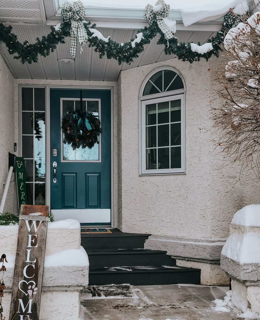 A wintery front porch decorated for Christmas, featuring a blue door, a wreath, garland with bows, and a wooden 'Welcome' sign. Snow is visible on the ground and roof.