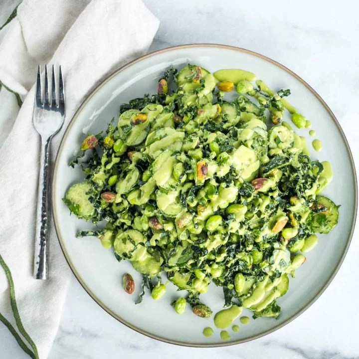 A plate of kale and cucumber salad topped with a creamy green dressing and sprinkled with pistachios, placed next to a fork on a marble surface.
