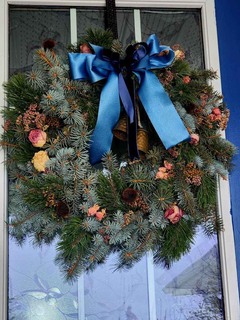 A beautifully decorated wreath made of pine branches, adorned with dried flowers, pine cones, and a large blue ribbon, hanging on a door.