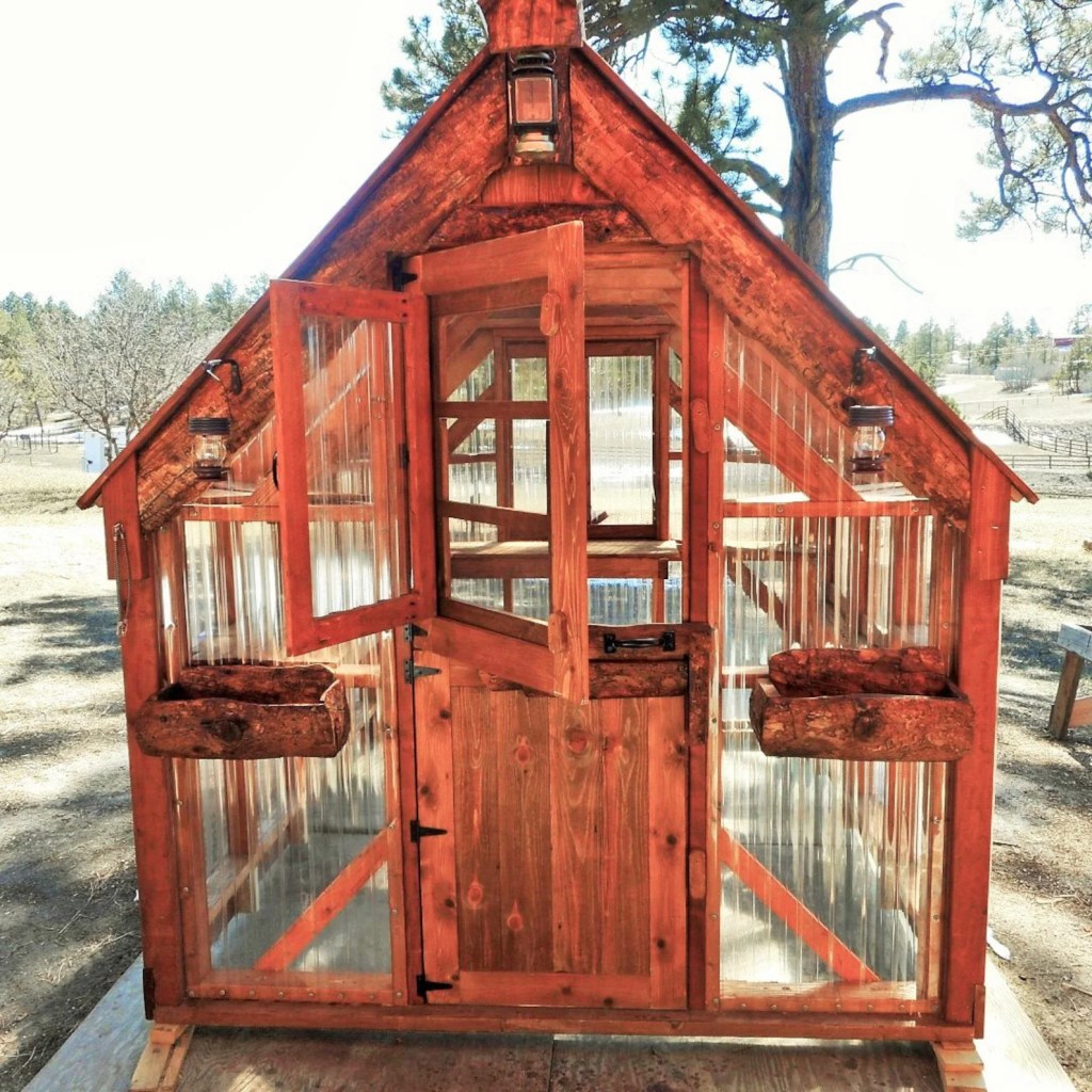 A wooden greenhouse with a peaked roof, featuring transparent panels, double doors, and hanging lanterns, set against a natural background.