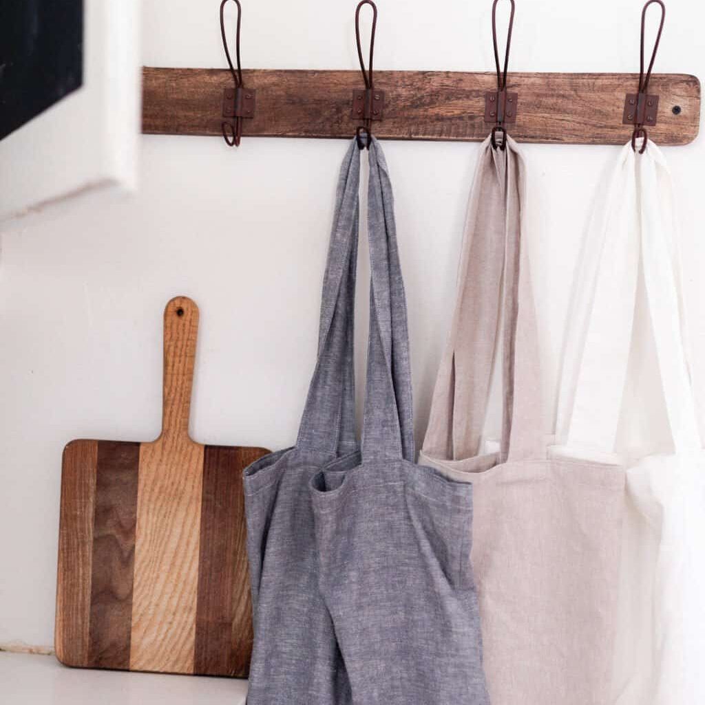 Three fabric tote bags hanging on a wooden hook rack, with a wooden cutting board placed on a surface nearby.