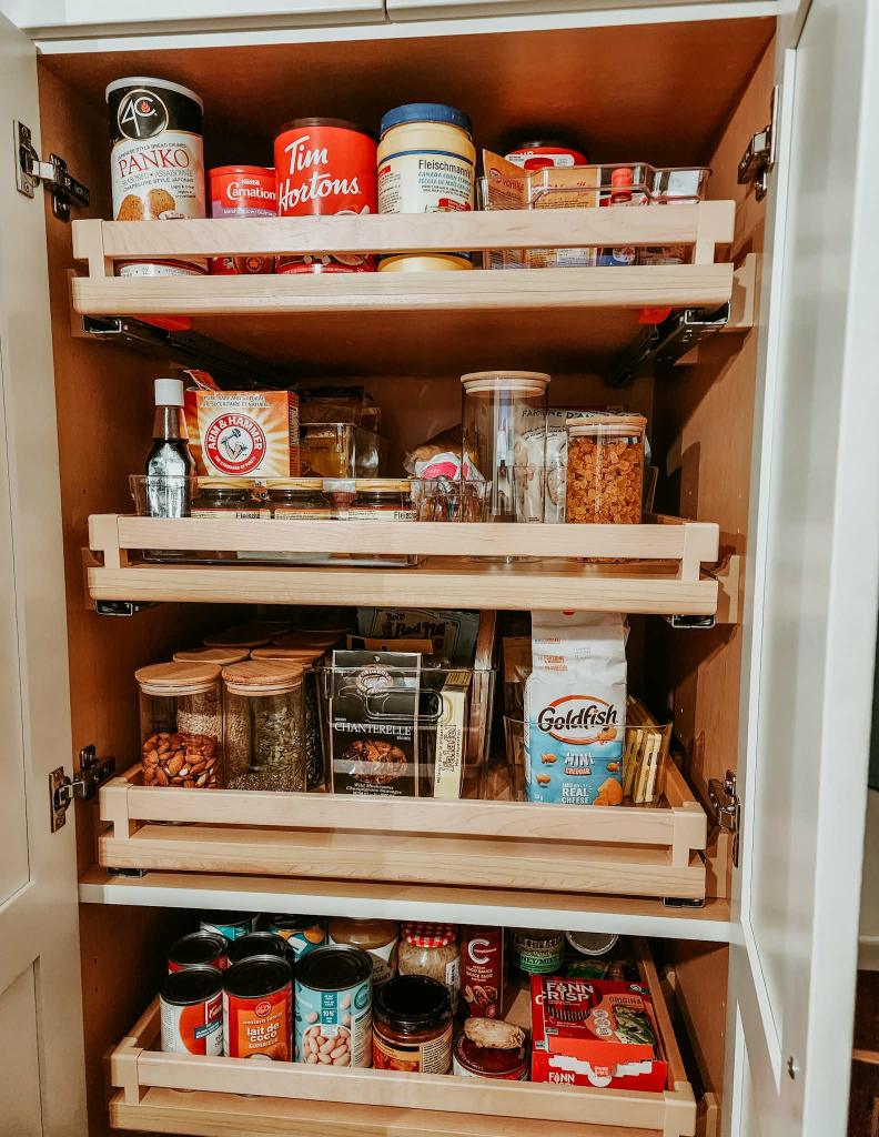 A wooden pantry cabinet with multiple shelves containing various food items, including canned goods, spices, baking supplies, and snacks. The pantry is organized with clear containers and boxes for easy access.