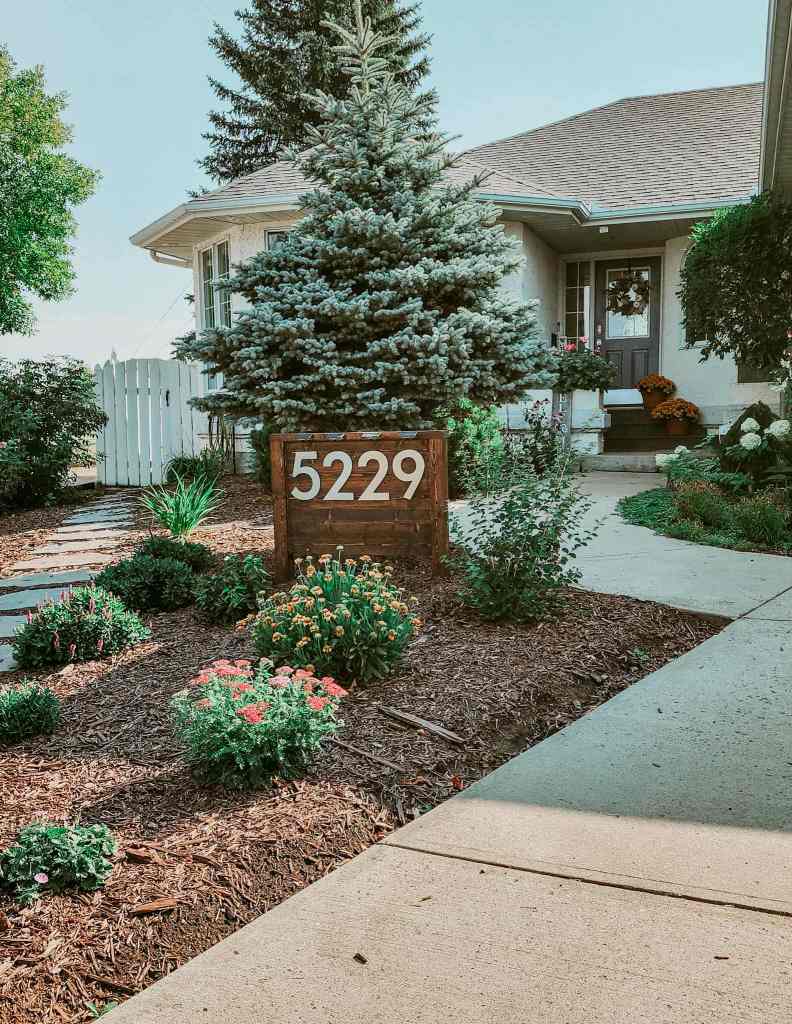 Front yard of a home featuring a wooden sign with the number '5229', surrounded by various plants and flowers, with a pathway leading to the entrance.