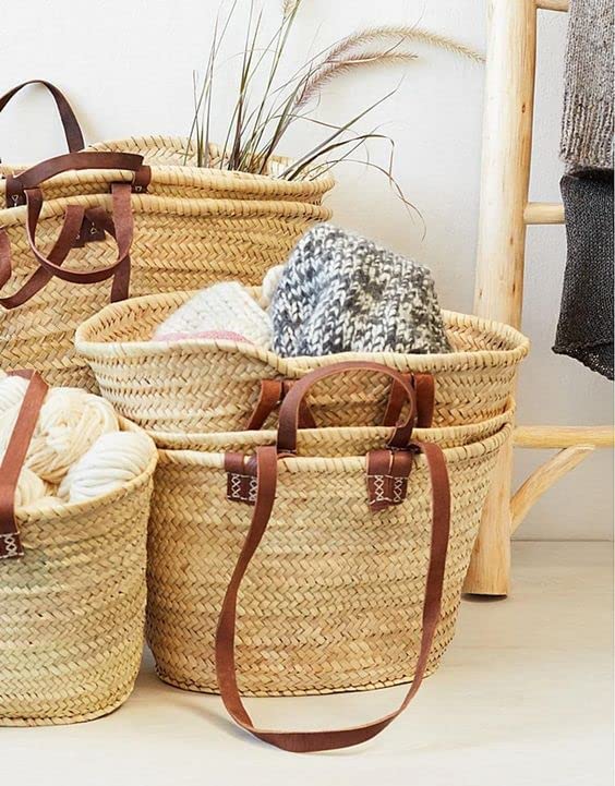 A collection of woven straw baskets with leather handles, filled with yarn and blankets, displayed against a light wall.