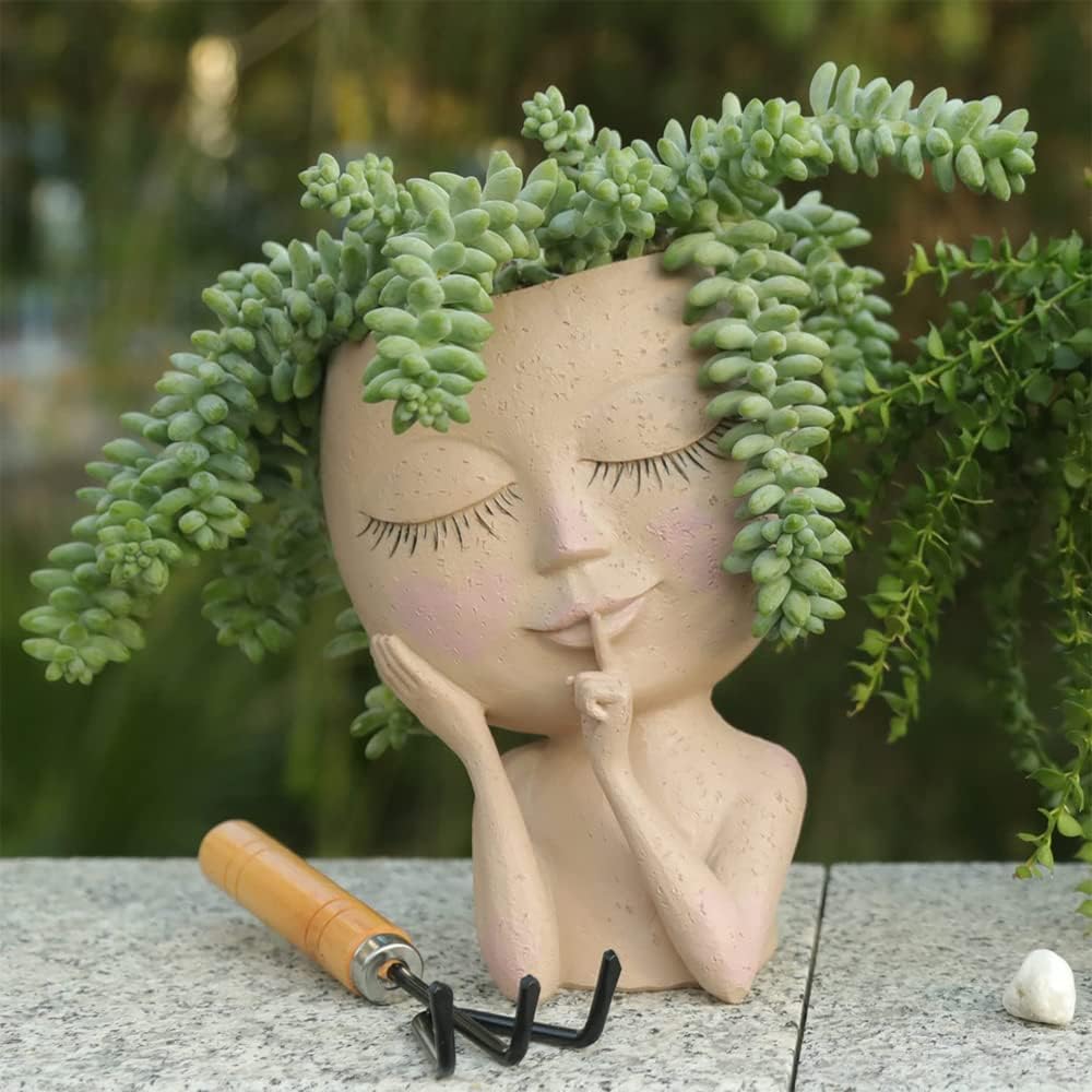 A whimsical planter shaped like a girl with closed eyes, adorned with green succulent plants as hair, resting on a stone surface beside a small gardening tool.
