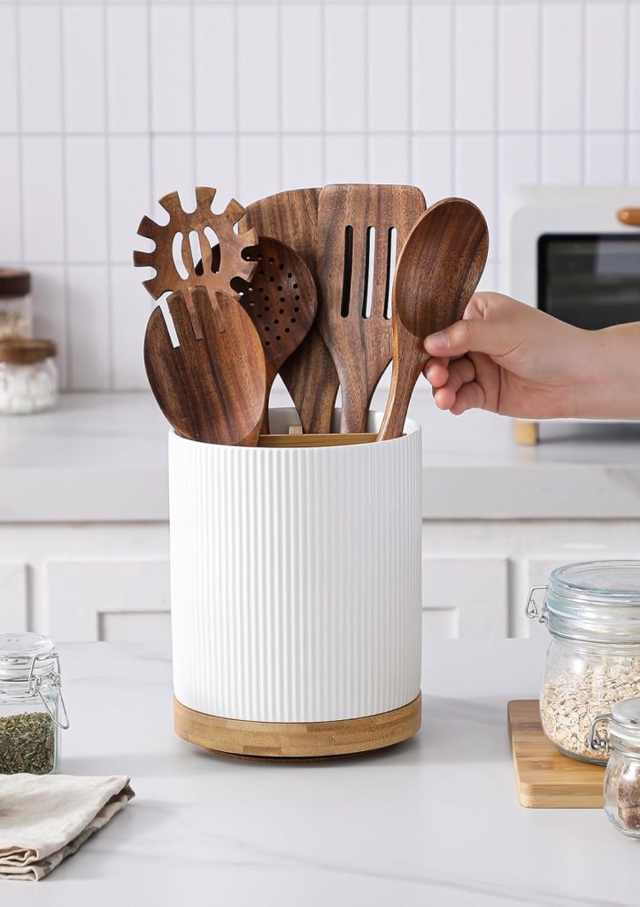 A hand reaching for a wooden kitchen utensil from a white, ribbed container on a marble countertop, surrounded by various kitchen items.