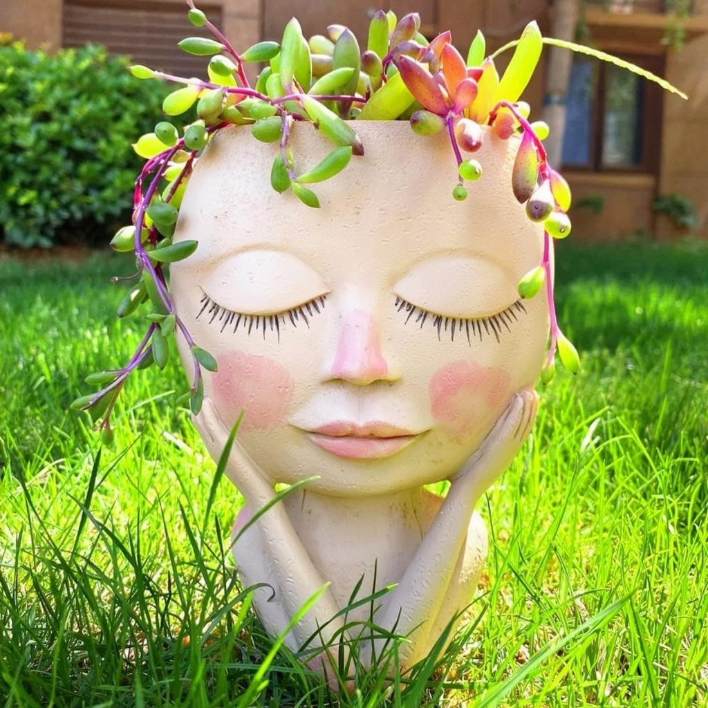 A decorative planter shaped like a serene face, featuring closed eyes and a gentle smile, adorned with various succulent plants resembling hair. Positioned on green grass.