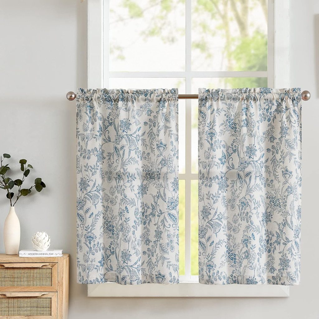 Close-up of light fabric curtains with a floral pattern in blue and white hanging by a window, with natural light filtering through.