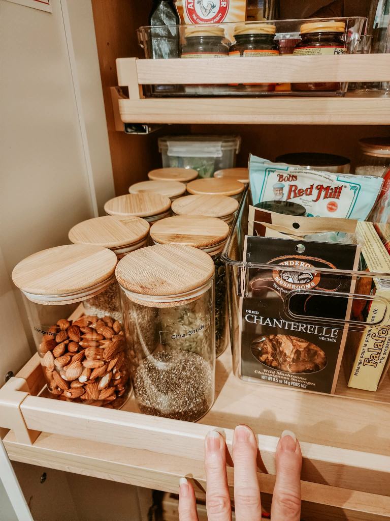 A kitchen pantry shelf displaying various storage jars with bamboo lids, containing almonds, chia seeds, and spices, along with a clear container of dried chanterelle mushrooms and other food items.
