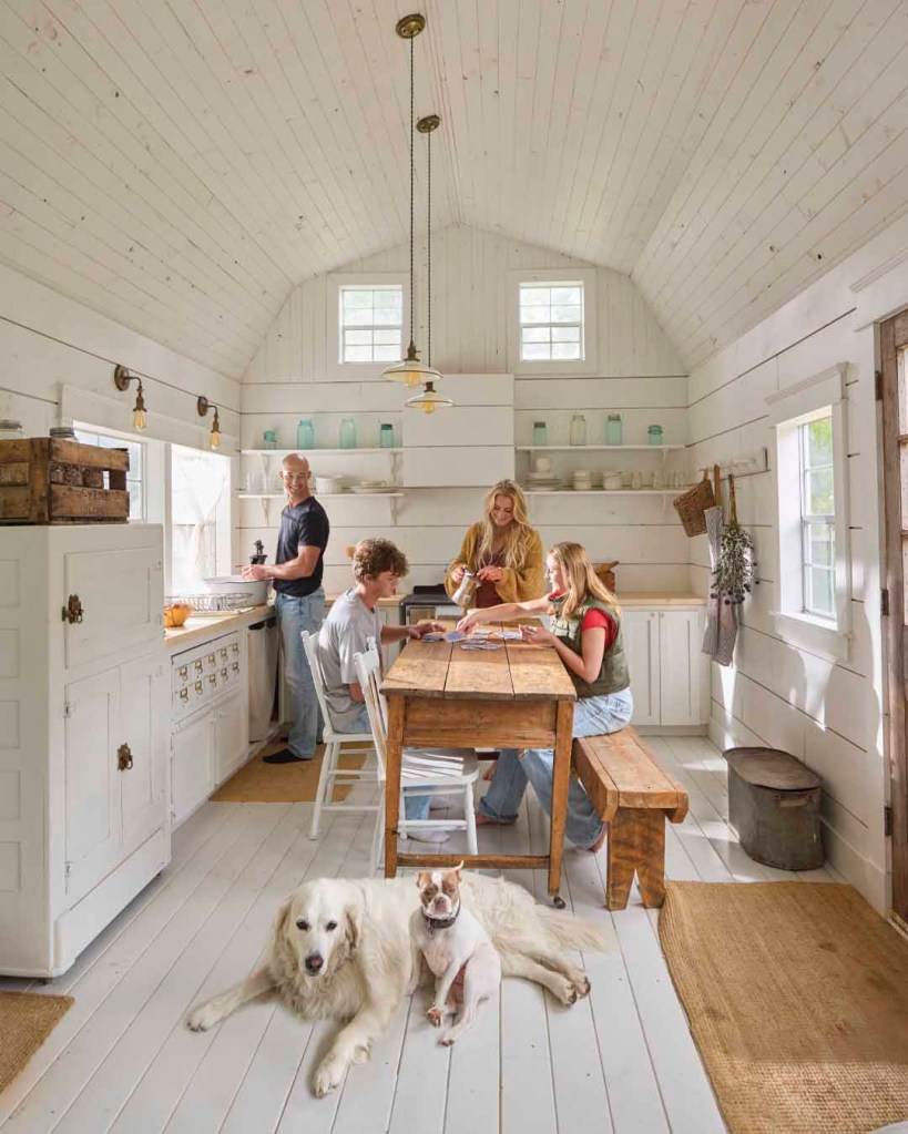 A cozy, white-walled kitchen scene featuring a family of four engaged in various activities around a wooden dining table, with two dogs resting on the floor.