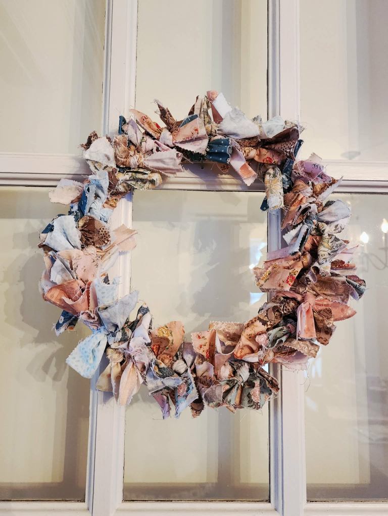 A handmade fabric wreath made of various patterned and textured fabric scraps, displayed on a window.