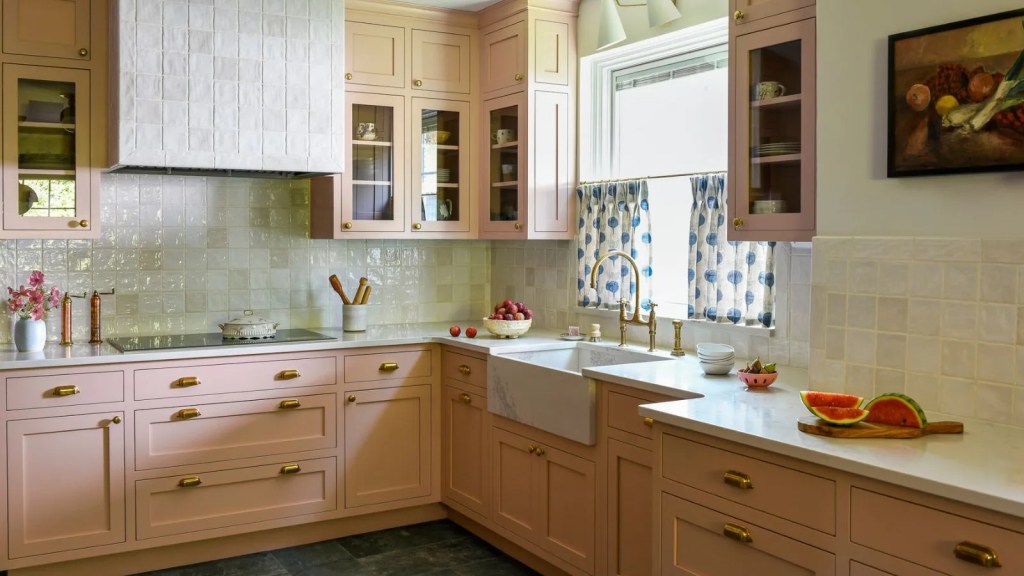 A beautifully designed kitchen featuring pink cabinetry, marble countertops, a farmhouse sink, and decorative window treatments, adorned with fresh flowers and fruit.