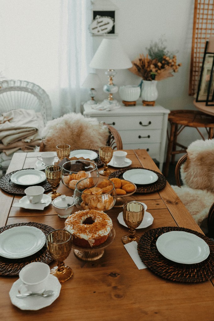 A beautifully arranged dining table featuring white plates, gold-rimmed glasses, and a variety of pastries including a large frosted donut and smaller donuts in glass bowls, set in a cozy and well-decorated room.