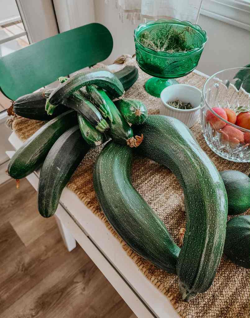 A collection of fresh zucchinis arranged on a wooden table, with a green chair and various bowls containing herbs and peaches in the background.
