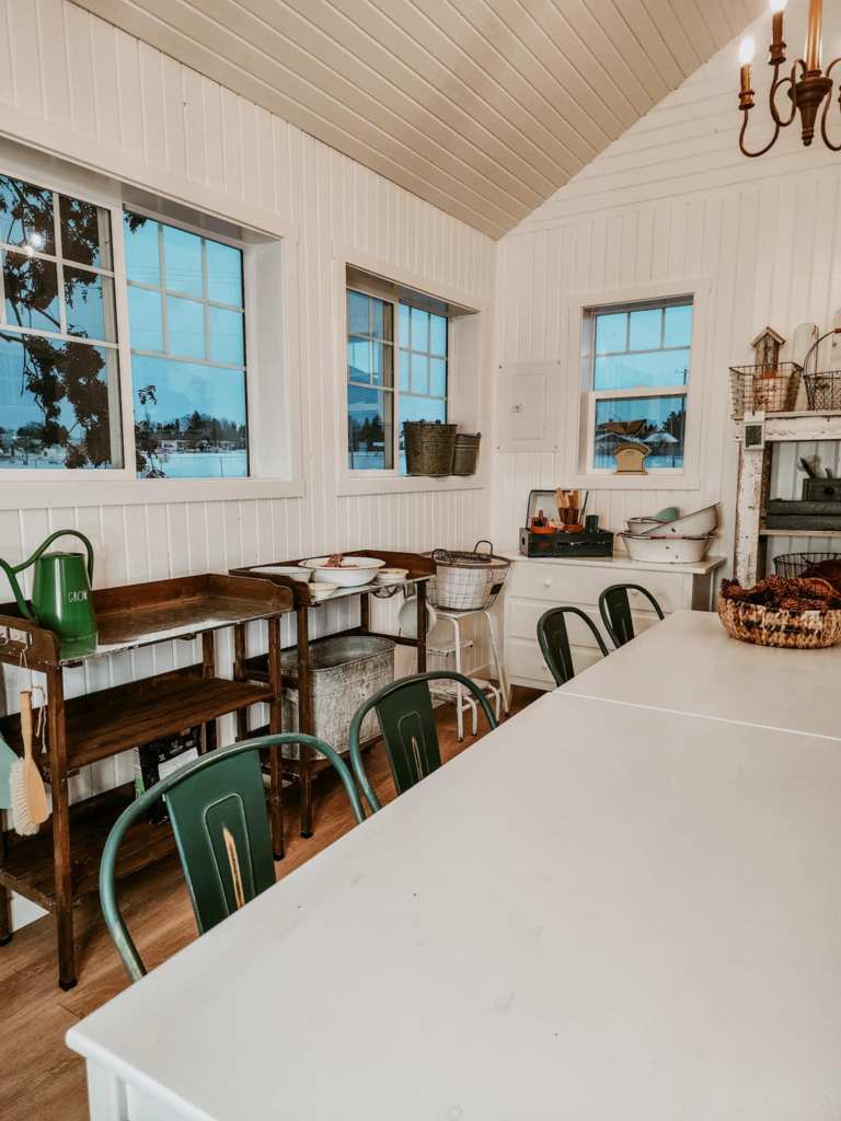 A bright, rustic kitchen interior featuring white wooden walls, a large table, green metal chairs, and windows allowing natural light in. A wooden shelf with various kitchenware and decorative baskets is visible.