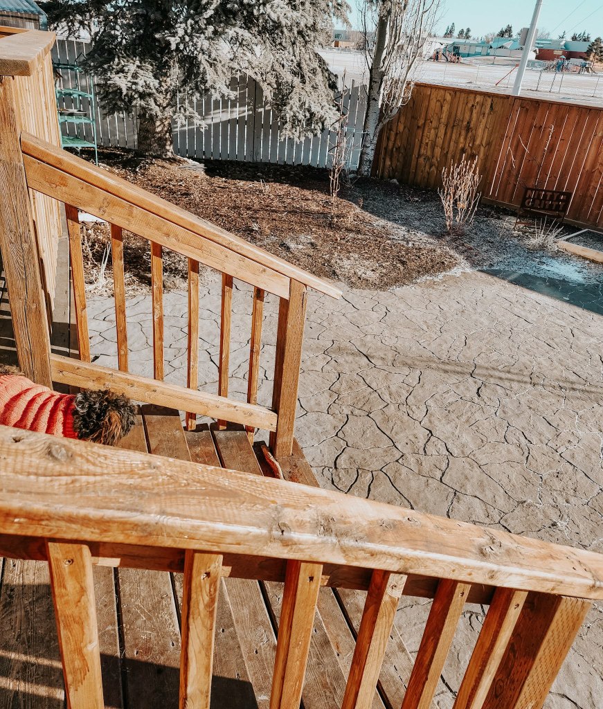View of a wooden staircase leading down to a landscaped yard with a patterned concrete path and a white fence in the background.