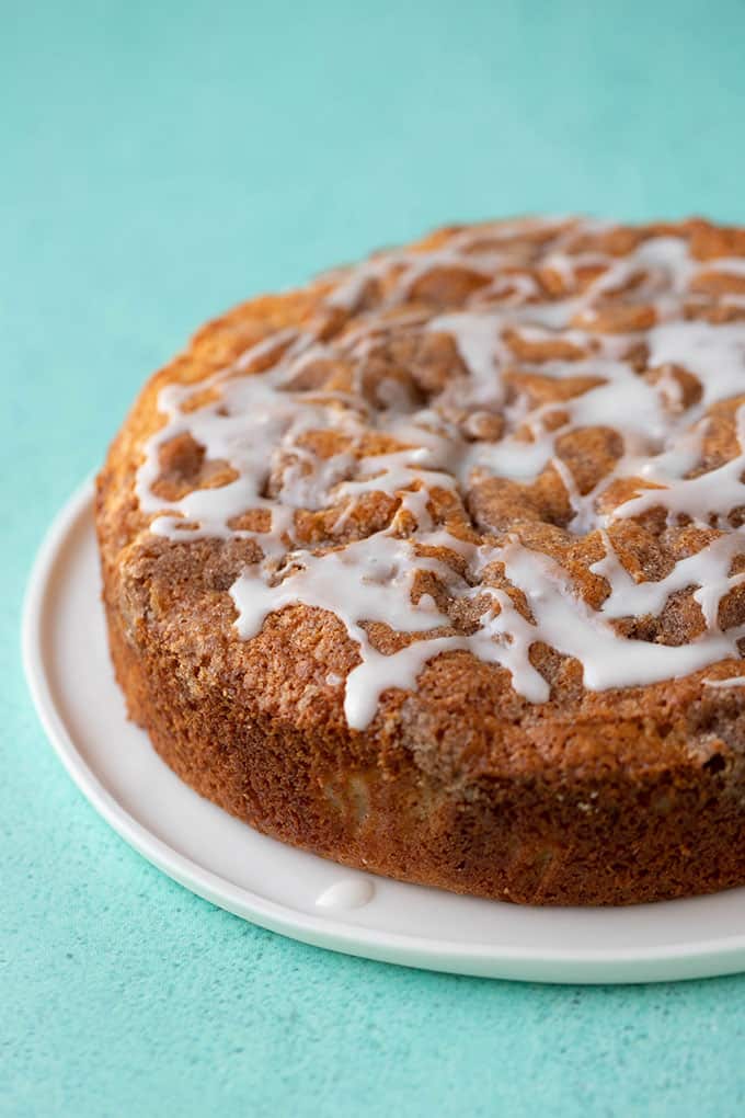 A close-up of an apple cake with a golden-brown crust, topped with a white icing drizzle, displayed on a white plate against a turquoise background.