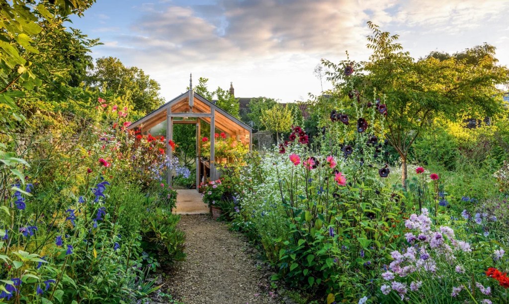 A vibrant garden path surrounded by colorful flowers leading to a wooden greenhouse under a partly cloudy sky.