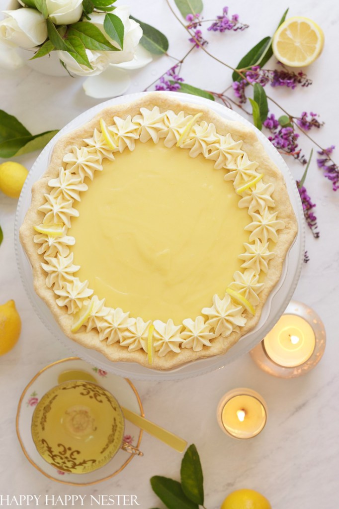 A beautifully decorated lemon pie with a smooth yellow filling, topped with star-shaped piped cream and fresh lemon slices, surrounded by lemon halves and floral arrangements on a white table.