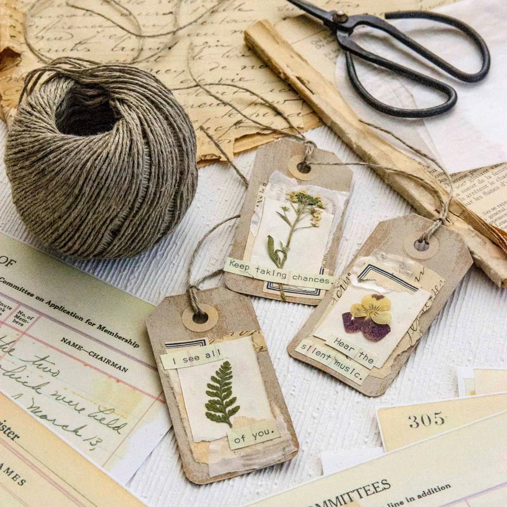 A collection of handcrafted tags with pressed flowers and inspirational phrases, arranged on a textured surface with spool of twine, scissors, and vintage papers.