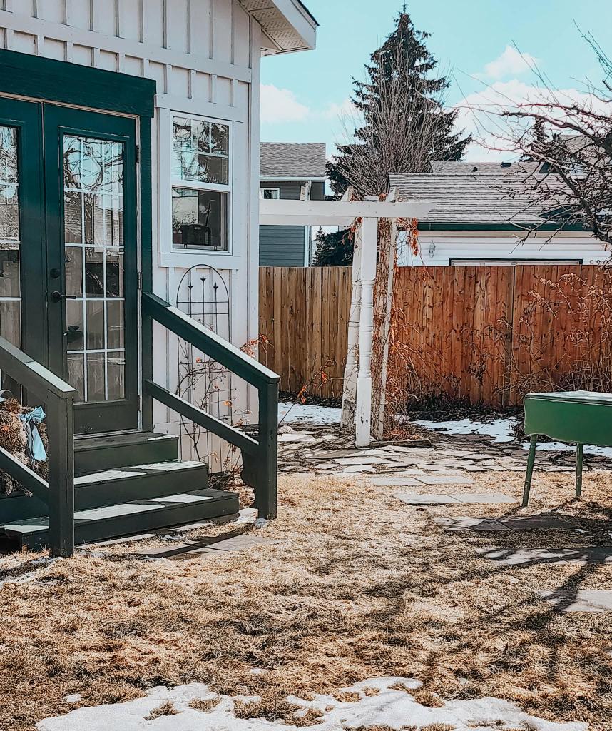 A backyard scene featuring a green door and steps leading up to a white house. There is a stone pathway in the foreground, a patch of brown grass, and some melting snow. A wooden fence and trees are visible in the background.