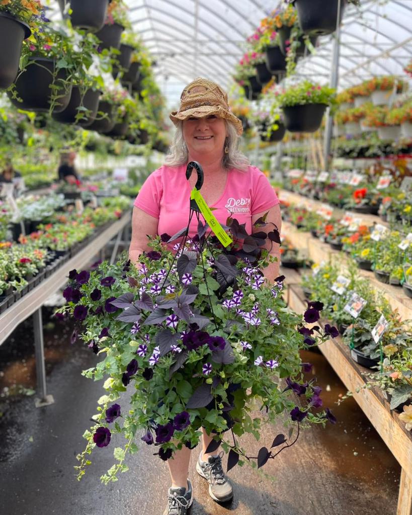 A smiling woman wearing a pink shirt and a straw hat stands in a greenhouse holding a large hanging basket filled with purple petunias and green plants, surrounded by various flowers and plants.