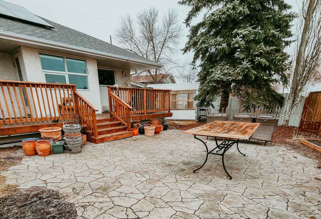 A backyard patio featuring a wooden deck, stone flooring, a dining table, and various potted plants, with a tall evergreen tree in the background.