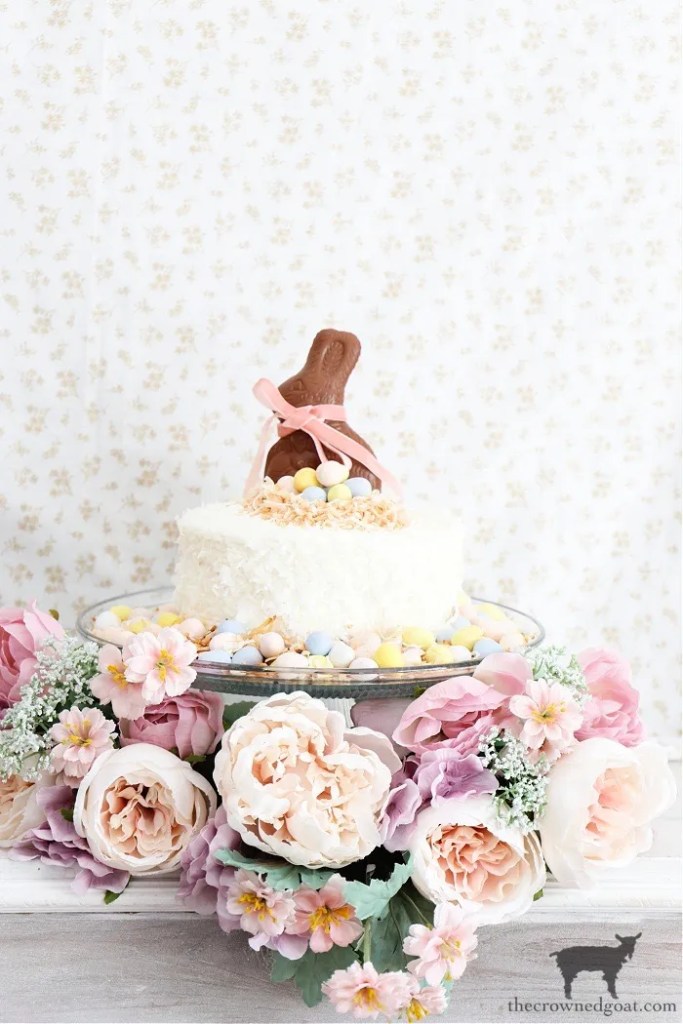 A decorated cake topped with a chocolate bunny and surrounded by pastel candy eggs, displayed on a glass cake stand with a floral arrangement of pink and white flowers.