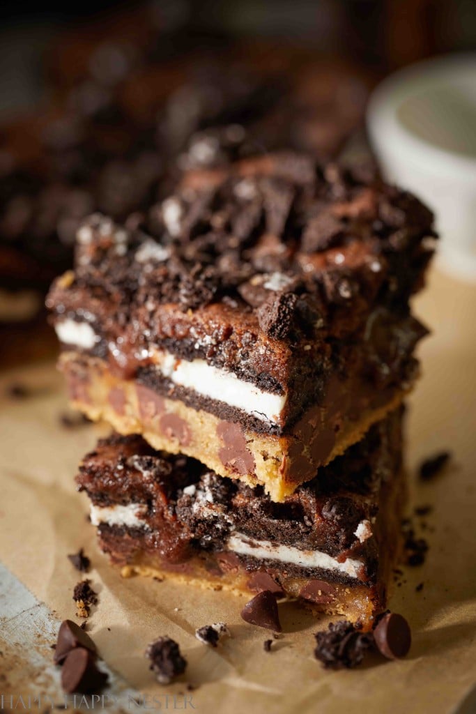 Layered chocolate dessert bars with chocolate chips, Oreo cookies, and cookie dough, stacked on parchment paper.
