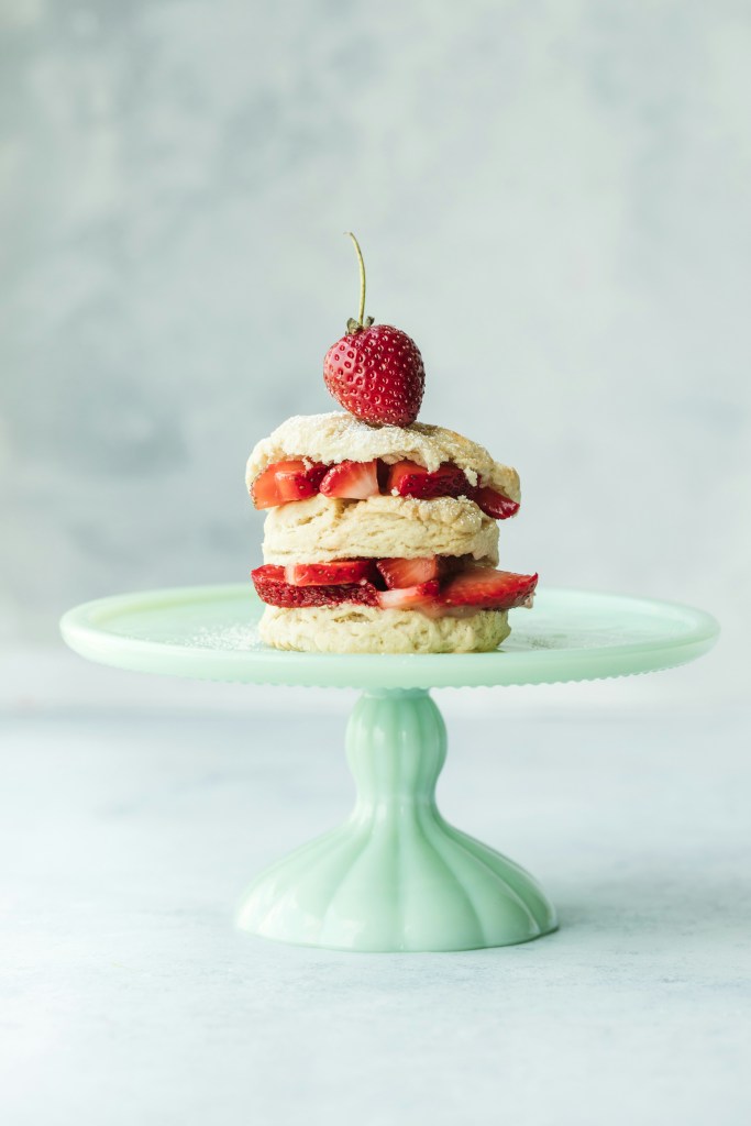 A layered strawberry shortcake topped with a fresh strawberry, presented on a mint green cake stand.