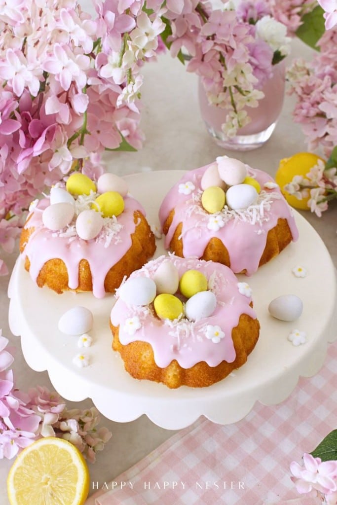 Three decorated mini bundt cakes with pink icing and colorful candy eggs, surrounded by pink flowers and a lemon slice.
