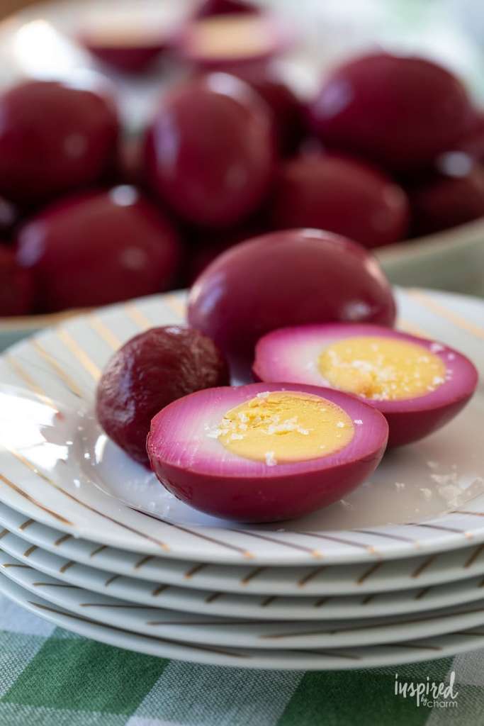 Sliced and whole pickled purple eggs on white plates with a striped design, with a green checkered tablecloth in the background.