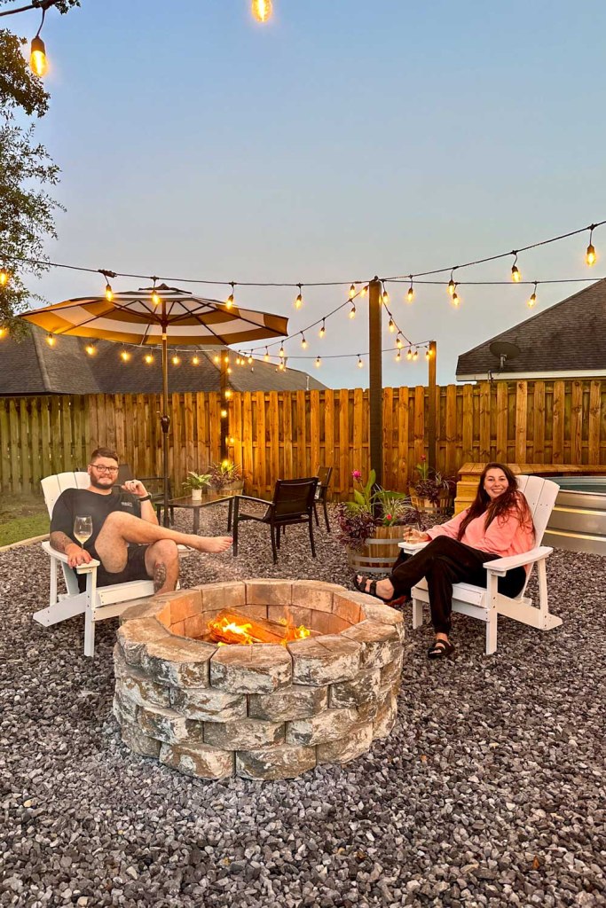 A couple sitting in Adirondack chairs in front of a fire pit, with string lights above.
