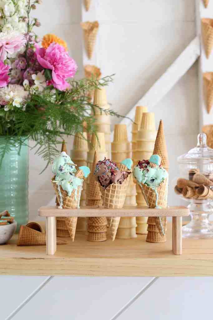 Three ice cream cones in a wooden ice cream cone holder sit on the table in front of some flowers and stacks of ice cream cones. 