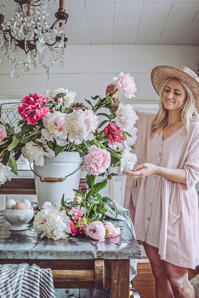 A lady in a pink dress making a bouquet of peonies in a white enamel pail.