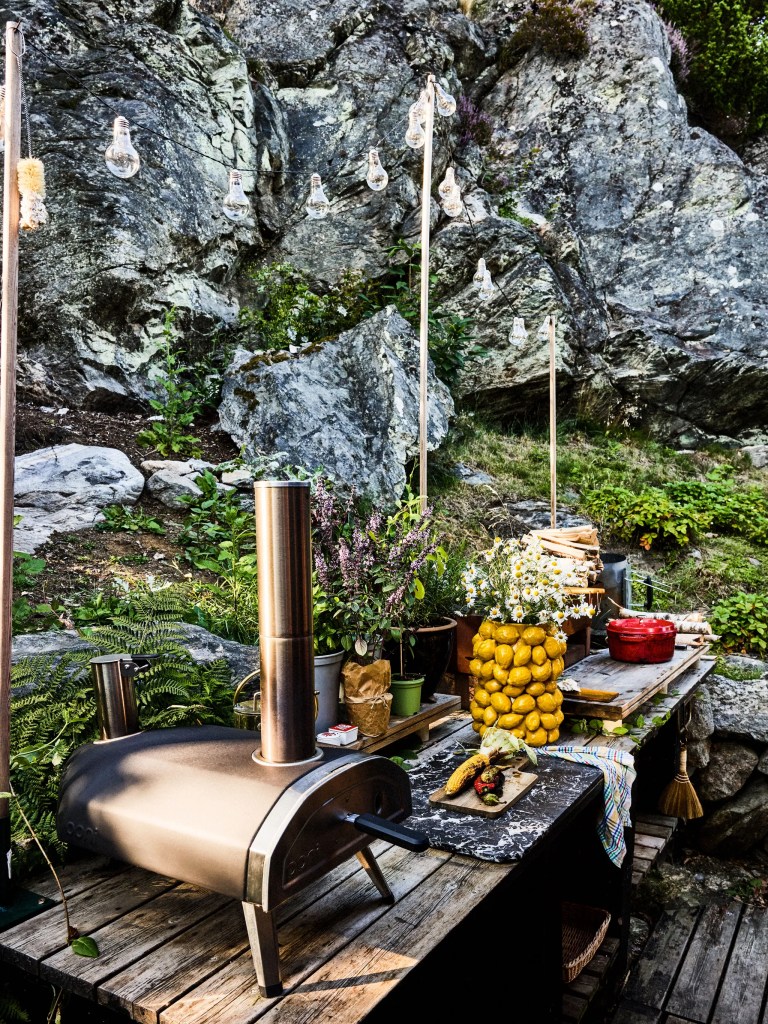 A pizza oven, table of herbs and flowers, in front of a grey cliff face.