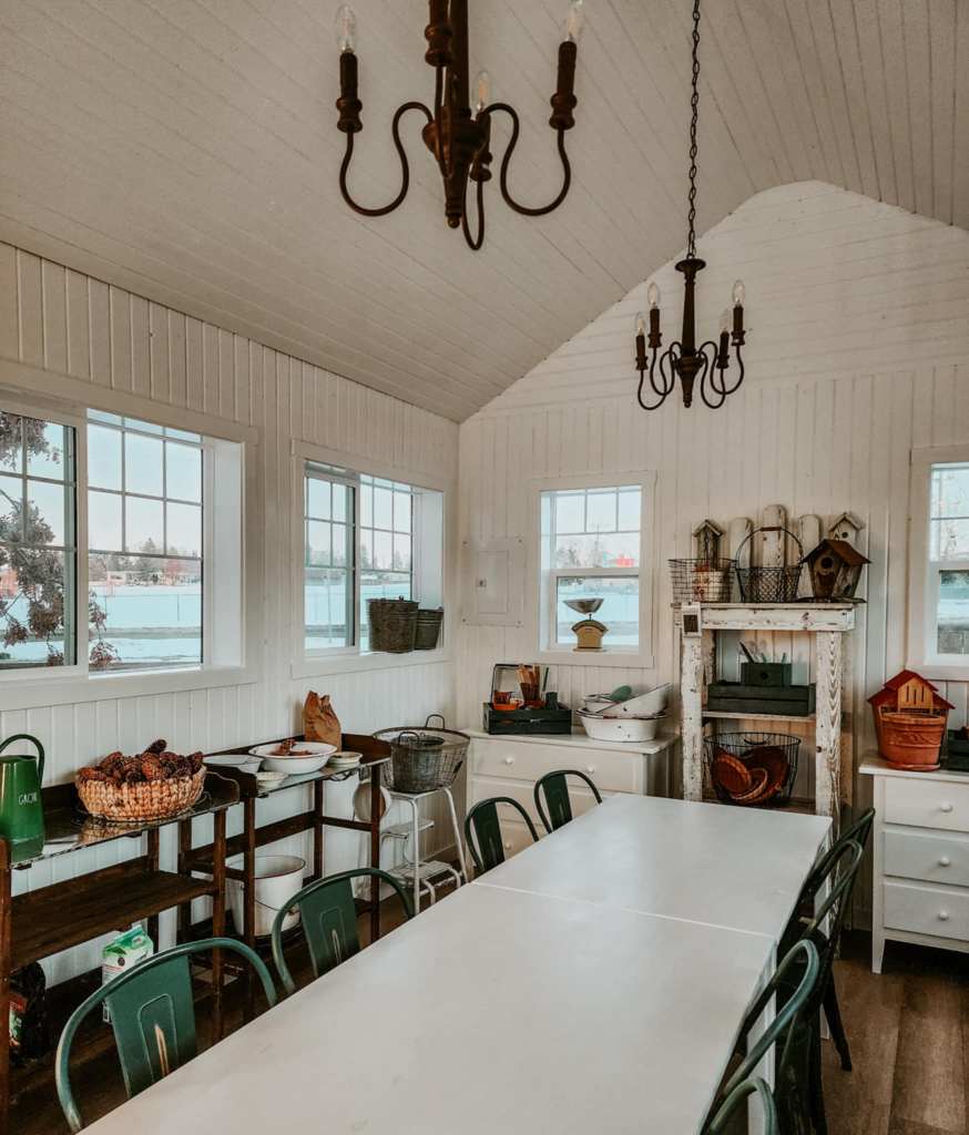 A garden shed with white walls, furniture and tables. Garden supplies on shelves and potting tables, forest green chairs, and antique chandelier's.
